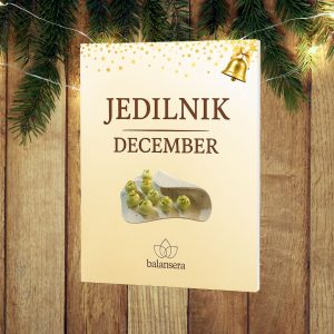 3D naslovnica decembrskega jedilnika Balansera, postavljena na lesenem prazničnem ozadju z vejicami iglavcev in toplimi lučkami.