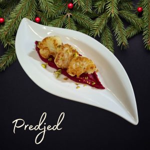 Praznična predjed z opečeno cvetačo na pesinem namazu, postrežena na elegantnem belem krožniku s smrečnimi vejicami v ozadju.